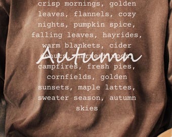 Puede incluir: Una camiseta marrón con la palabra "Autumn" en escritura blanca, que enumera palabras con temática otoñal. La persona está sentada en un sofá de color claro, sosteniendo una taza blanca. El texto de la camisa incluye "mañanas frescas" y "hojas doradas."