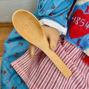 May include: A light-colored wooden spoon with a rounded bowl and a long handle. The spoon is held over a red and white striped fabric and a blue fabric with a floral pattern. The image also shows a red heart with the number "1854" stitched on it.