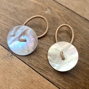 May include: Two iridescent button hair ties with elastic bands. The buttons are round and have a pearlescent sheen, reflecting colors like white, pink, and blue. The elastic bands are a light brown color.