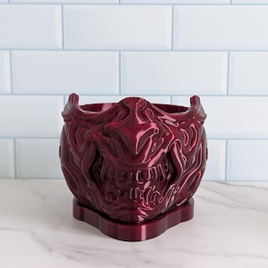 May include: A burgundy-colored decorative planter shaped like a Japanese Oni mask. The planter has a detailed, textured surface with a wide opening for plants. The base is rectangular, providing stability.