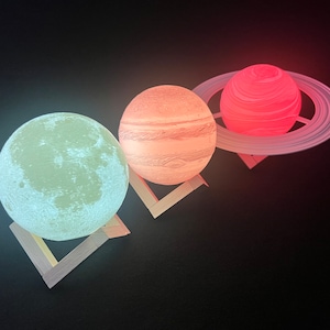 May include: Three illuminated planet-shaped lamps: a moon, Jupiter, and Saturn. The moon glows pale green, Jupiter is orange with stripes, and Saturn is red with a ring. Each lamp sits on a small wooden stand against a black background.