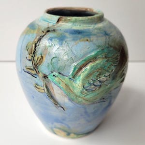 Peut inclure: Un vase en céramique au corps arrondi et à l'ouverture étroite, présentant une glaçure bleu clair avec des nuances tourbillonnantes de blanc et de vert. Un relief texturé d'un oiseau en vol tenant une branche est centré sur le devant.