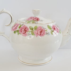 May include: A white ceramic teapot with a lid, handle, and spout. The teapot is decorated with pink roses and green leaves. Gold trim accents the handle, spout, and base. A classic design for tea.