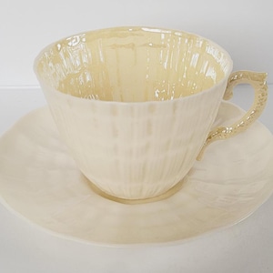 May include: A cream-coloured teacup and saucer set. The teacup has a textured surface and a decorative handle. The saucer is round with a matching textured design. The set is made of ceramic.