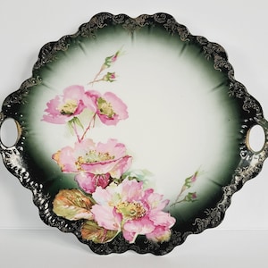 May include: An antique serving tray with a scalloped edge and handles. The tray features a hand-painted floral design of pink roses with green leaves against a white and dark green background. The rim is detailed with a silver filigree.