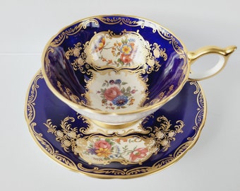 May include: A vintage teacup and saucer set. The cup and saucer are a deep blue with gold trim and floral designs. The center of the cup and saucer is white with a floral pattern. The handle of the cup is gold.
