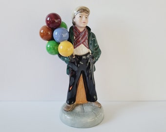 May include: A ceramic figurine of a young boy holding a bunch of colorful balloons. The boy wears a green jacket, black pants, and a cap. The balloons are various colors, including red, brown, blue, green, and yellow.