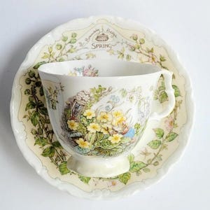 May include: A porcelain teacup and saucer set featuring the Brambly Hedge Spring design. The cream-coloured pieces are decorated with illustrations of yellow primrose flowers, green foliage, and small animal characters. The saucer includes the text SPRING.