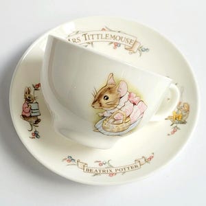 May include: A cream-colored ceramic teacup and saucer set featuring illustrations of Mrs. Tittlemouse. The cup displays a mouse in a pink dress with a basket, while the saucer includes the text Mrs. Tittlemouse and Beatrix Potter with floral accents.