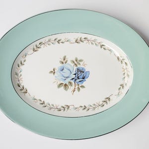 May include: An oval serving platter featuring a wide mint green border with a silver rim. The centre is white, decorated with a central cluster of two blue roses and a surrounding delicate wreath of small green leaves and tiny blue flowers.