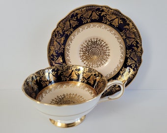 May include: A vintage teacup, saucer, and plate set. The teacup and saucer feature a cream-colored interior with a gold floral design. The exterior of the teacup and the plate are navy blue with gold floral patterns. The set has gold trim.