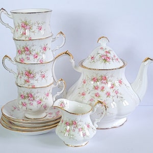 Peut inclure: Service à thé en porcelaine blanche comprenant une théière, un pot à crème et des tasses empilées avec des soucoupes. L'ensemble est orné de roses roses et de bordures dorées. La théière a un bouton en forme de coquillage.