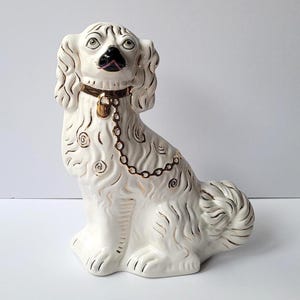 以下が含まれることがあります： 白い陶器製の犬の置物。毛並みに金色のアクセントが施され、小さな南京錠が付いた金色の首輪と、胸元に垂れ下がった金色のチェーンが特徴です。黒い鼻と大きな目をした犬が、座った姿勢で描かれています。