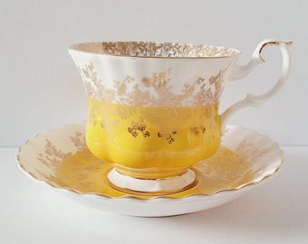 May include: A vintage teacup and saucer set. The teacup is white with gold filigree and a bright yellow band. The saucer is white with gold trim. The handle of the teacup is curved. A classic tea set for afternoon tea.