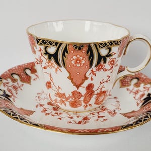 May include: A decorative teacup and saucer set. The cup and saucer have a white base with a detailed coral, black, and gold pattern. The cup has a curved handle and a gold rim, and the saucer has a matching design.