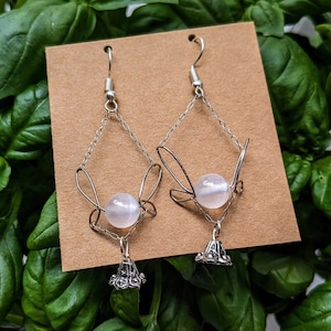 Puede incluir: Un par de pendientes plateados con un diseño geométrico. Cada pendiente presenta una cuenta redonda y transparente, una cadena y un dije decorativo en forma de campana. Los pendientes se exhiben sobre una tarjeta marrón.