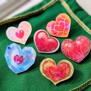 Könnte beinhalten: Eine Sammlung von sechs herzförmigen Anstecknadeln mit verschiedenen Designs. Die Anstecknadeln weisen Farben wie Rosa, Rot, Orange und Blau auf, mit Mustern wie Wirbeln und geometrischen Formen. Sie sind auf einem grünen Stoff angeordnet.