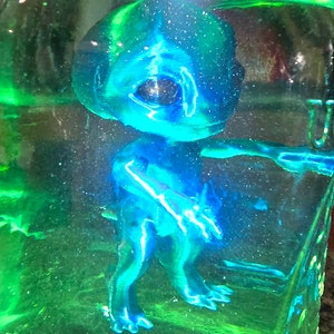May include: A blue alien figurine inside a clear, rectangular container filled with a green liquid. The alien has large eyes and outstretched arms. The container has a green tint.