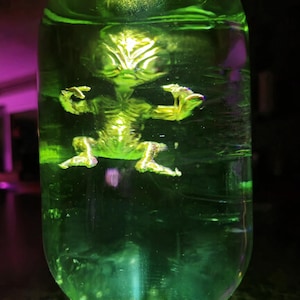 May include: A clear glass bottle filled with a vibrant green liquid, housing a small, golden alien figurine. The alien is posed with arms outstretched. The bottle's contents are illuminated, creating a glowing effect.