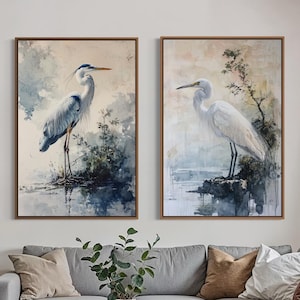 Pode incluir: Duas impressões de arte emolduradas com pinturas em aquarela de garças em um ambiente natural sereno. A obra de arte apresenta uma garça azul e uma garça branca, cada uma em pé na água. As impressões são exibidas acima de um sofá seccional cinza em uma sala de estar.