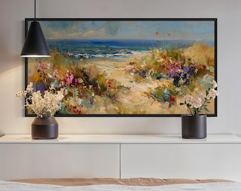 Puede incluir: Un cuadro al óleo rectangular enmarcado que representa un paisaje costero con un sendero de arena, flores silvestres coloridas y un océano azul bajo un cielo nublado. La obra se exhibe sobre un mueble blanco con dos jarrones oscuros con flores.