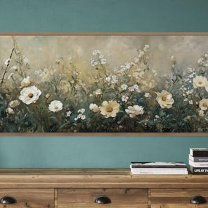 May include: A framed floral painting in neutral tones of beige, cream, and green. The artwork shows a field of white and yellow flowers. A clear glass vase with branches is on the left. A wooden cabinet with books is below.