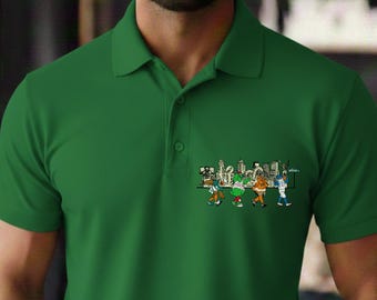 May include: A green polo shirt with a small cartoon graphic on the chest pocket. The graphic depicts a line of cartoon characters in front of a city skyline. The shirt has a collared neckline and short sleeves.