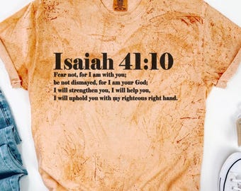 May include: Peach-colored t-shirt with the text "Isaiah 41:10" and a Bible verse printed in black. The shirt is paired with blue jeans and white sneakers. The shirt has a unique, textured design.