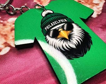 May include: Green keychain in the shape of a t-shirt. The keychain features a graphic of an eagle wearing sunglasses and a green beanie with the word "PHILADELPHIA" on it. The keychain is attached to a silver metal keyring.