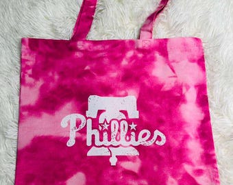 May include: A pink tie-dye tote bag with the Philadelphia Phillies logo in white. The bag has a rectangular shape with a matching pink handle. The Phillies logo features the team name and a bell symbol.