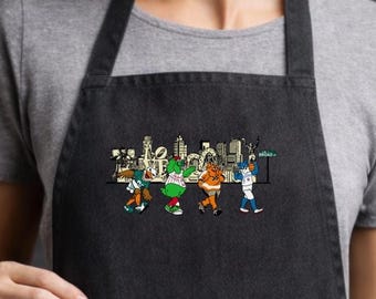 May include: Black denim apron with a colorful embroidered design of cartoon figures in front of a city skyline. The apron is worn by a person in a gray t-shirt, with a kitchen setting in the background.