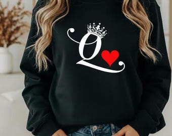 May include: Black sweatshirt with a white "Q" design, a crown, and a red heart. The "Q" is stylized with a decorative flourish. The person is wearing blue jeans.