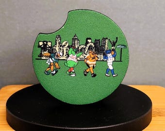 May include: Green fabric car coaster featuring a cartoon cityscape and four cartoon characters. The coaster has a curved edge for easy placement in a car cup holder. The characters are in various sports uniforms.