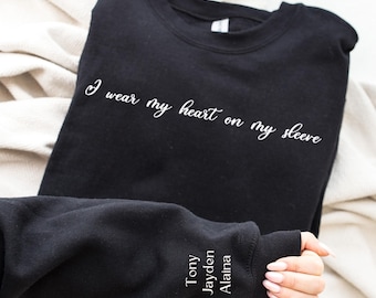 May include: Black sweatshirt with the text "I wear my heart on my sleeve" printed on the front. The sleeve has the names "Tony, Jayden, Alaina" embroidered on it.