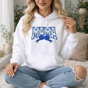 Può includere: Felpa con cappuccio bianca con tasca frontale e la scritta "MAMA" in stampa floreale blu. Un disegno a fiocco blu è centrato sotto la parola. La felpa è indossata con jeans blu.