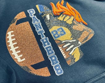 May include: Navy blue hooded sweatshirt featuring a football and a football player graphic. The football is glittery brown, and the player has orange hair and the number 23. The word "PANTHERS" is printed vertically.