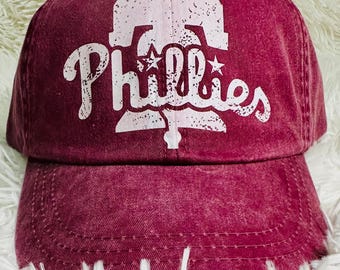 May include: A maroon baseball cap with the word "Phillies" in white script and a white graphic of the Liberty Bell. The cap has a curved brim and a distressed, vintage look. The hat is made of a soft, textured fabric.