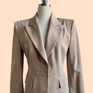 Pu&ograve; includere: Un blazer beige con revers a lancia, due tasche anteriori e chiusura a un bottone. La giacca su misura &egrave; esposta su un manichino su sfondo color pesca. Il blazer ha un design classico e professionale.