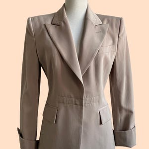 Pu&ograve; includere: Un blazer beige con revers a lancia, due tasche anteriori e polsini ripiegati. La giacca su misura &egrave; esposta su un manichino. Il blazer ha una chiusura a un bottone e uno stile classico e professionale.