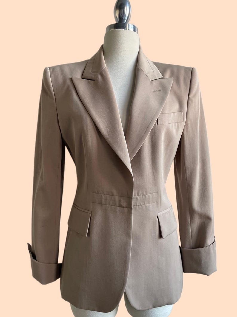 Pu&ograve; includere: Un blazer beige con revers a lancia, due tasche anteriori e polsini ripiegati. La giacca su misura &egrave; esposta su un manichino. Il blazer ha una chiusura a un bottone e uno stile classico e professionale.