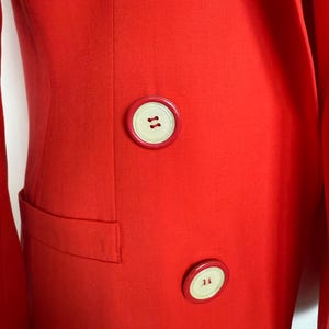 May include: A close-up of a bright red blazer with a tailored fit. The blazer features large, round, cream-colored buttons with brown stitching. A small pocket is visible on the left side. The fabric has a subtle texture.