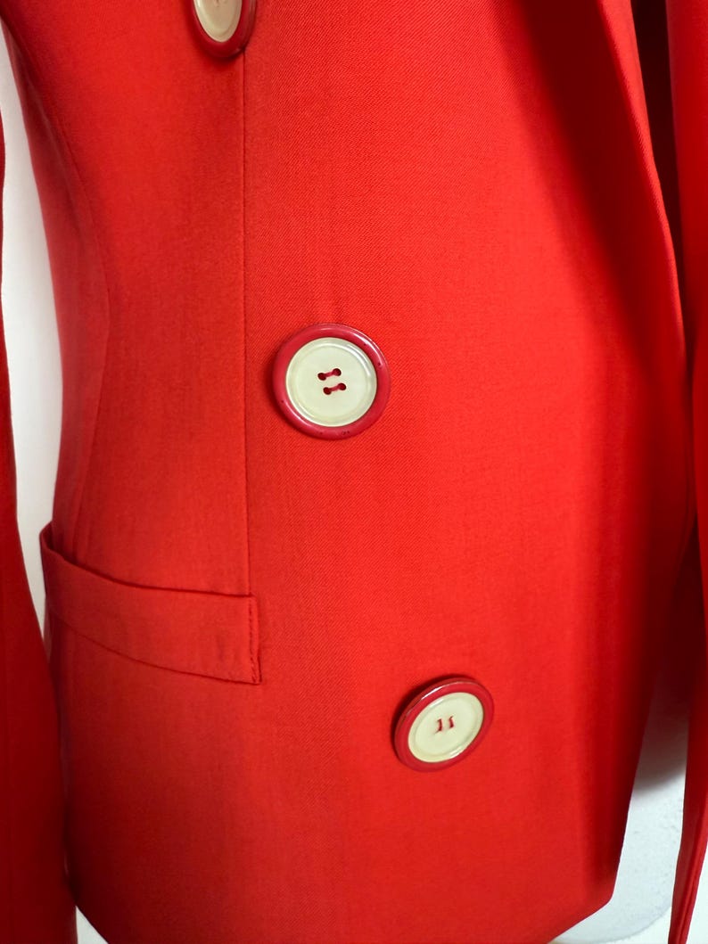 May include: A close-up of a bright red blazer with a tailored fit. The blazer features large, round, cream-colored buttons with brown stitching. A small pocket is visible on the left side. The fabric has a subtle texture.