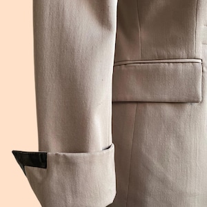 Pu&ograve; includere: Primo piano di una manica di blazer beige con un dettaglio nero sul polsino. Il blazer ha una vestibilit&agrave; sartoriale e un dettaglio tasca. Il tessuto sembra essere un materiale liscio e tessuto.