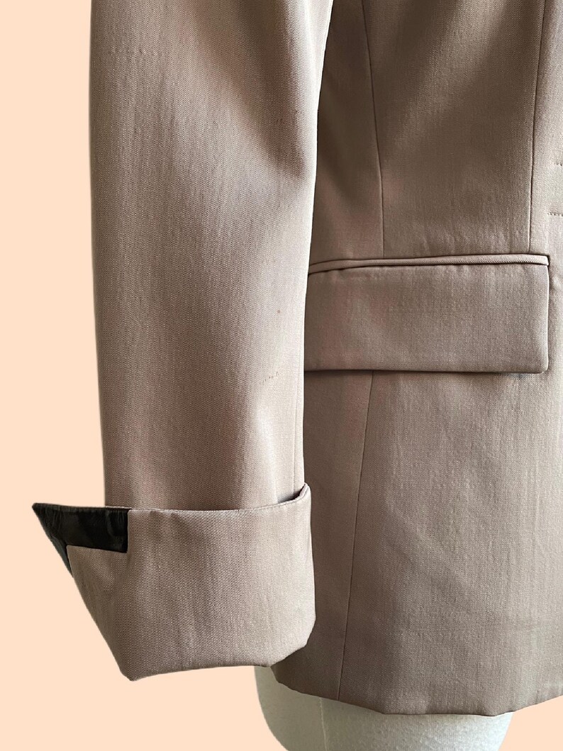 Pu&ograve; includere: Primo piano di una manica di blazer beige con un dettaglio nero sul polsino. Il blazer ha una vestibilit&agrave; sartoriale e un dettaglio tasca. Il tessuto sembra essere un materiale liscio e tessuto.