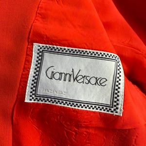 May include: Close-up of the interior of a bright orange garment, revealing a white label with the text "Gianni Versace" in black script. The label is framed with a black and white checkered border, set against a textured orange lining.