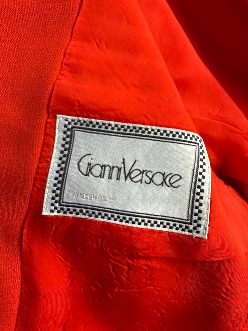 May include: Close-up of the interior of a bright orange garment, revealing a white label with the text "Gianni Versace" in black script. The label is framed with a black and white checkered border, set against a textured orange lining.