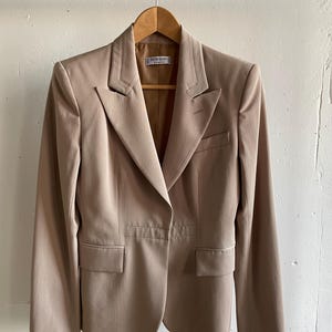 Pu&ograve; includere: Un blazer beige con revers a lancia, due tasche frontali e chiusura a un bottone. La giacca &egrave; appesa a una gruccia di legno. Il blazer ha una vestibilit&agrave; sartoriale e un design classico.