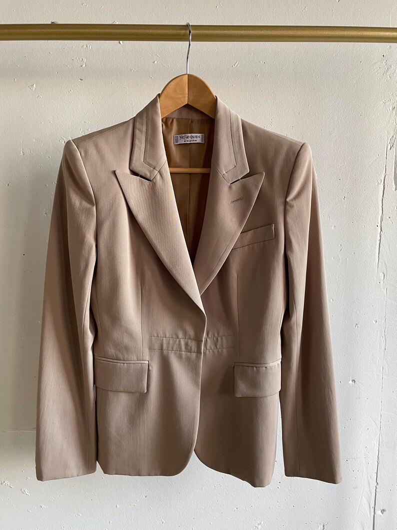 Pu&ograve; includere: Un blazer beige con revers a lancia, due tasche frontali e chiusura a un bottone. La giacca &egrave; appesa a una gruccia di legno. Il blazer ha una vestibilit&agrave; sartoriale e un design classico.