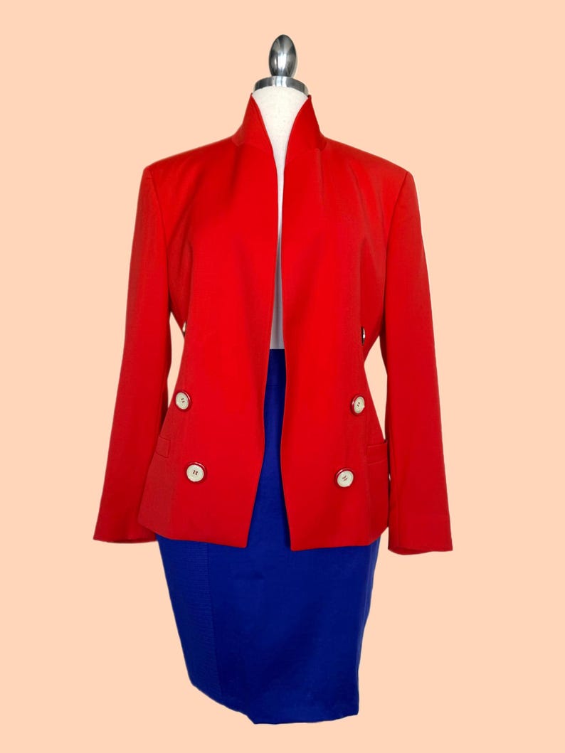 May include: A vibrant red blazer with a unique collar design and four white buttons. The blazer is paired with a royal blue skirt, creating a bold color contrast. The outfit is displayed on a mannequin.