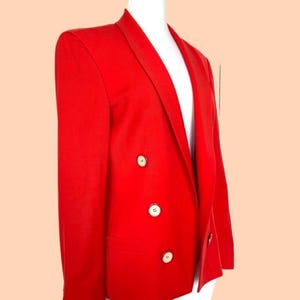 May include: A vibrant red blazer with a classic double-breasted design. The blazer features three white buttons on each side and a notched lapel. The tailored fit and bold color make it a statement piece. The blazer is displayed on a white mannequin.